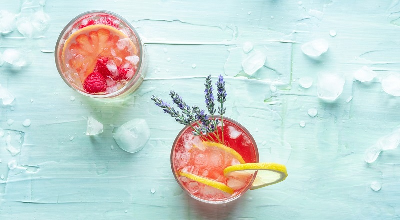 Two glasses of berry lemonade on a blue table with ice and lemon slices.