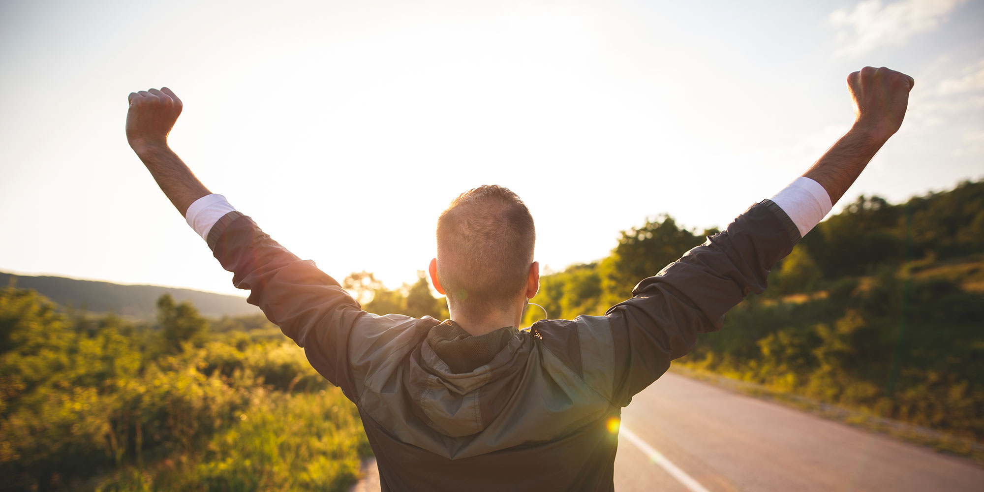 A man raises his arms in joyous celebration. He's standing on an empty road as the sun sets in the foreground.