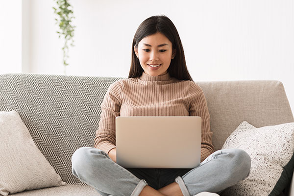 A young woman smirks as she types on her laptop. She's sitting on a gray couch.