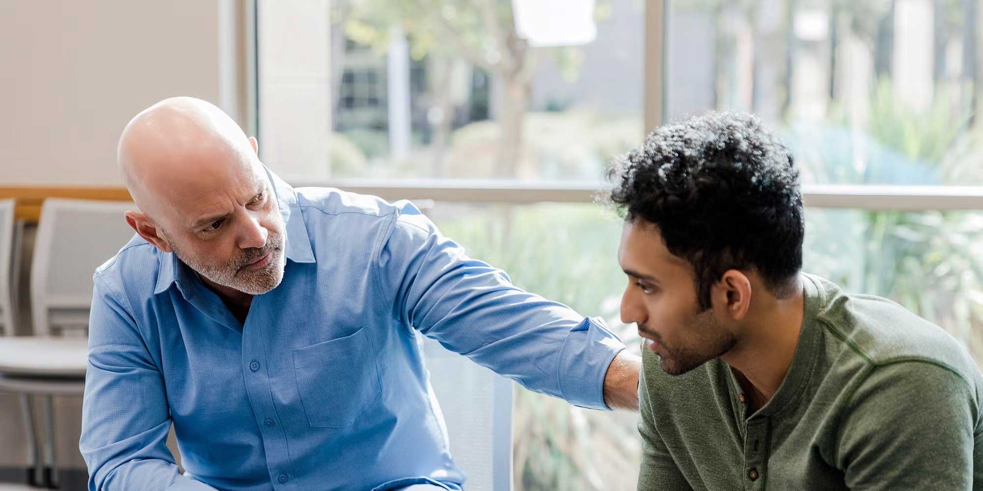 A middle-aged provider wearing a blue shirt sits leaning over, consoling his younger male patient