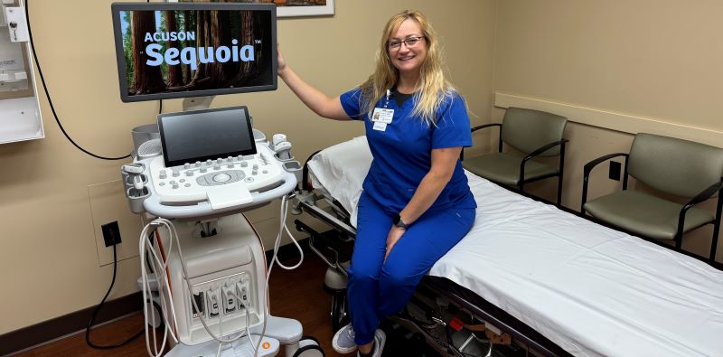 Amber sits in exam room wearing scrubs next to an ultrasound machine, smiling.