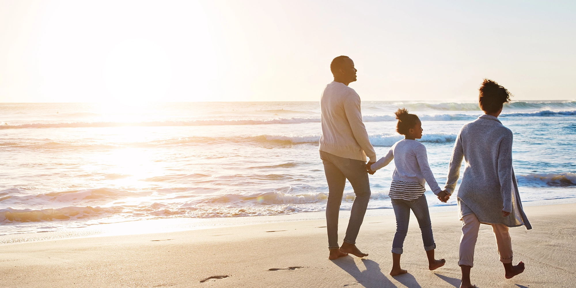 A family of three holds hand at the edge of the beach shore during sunset