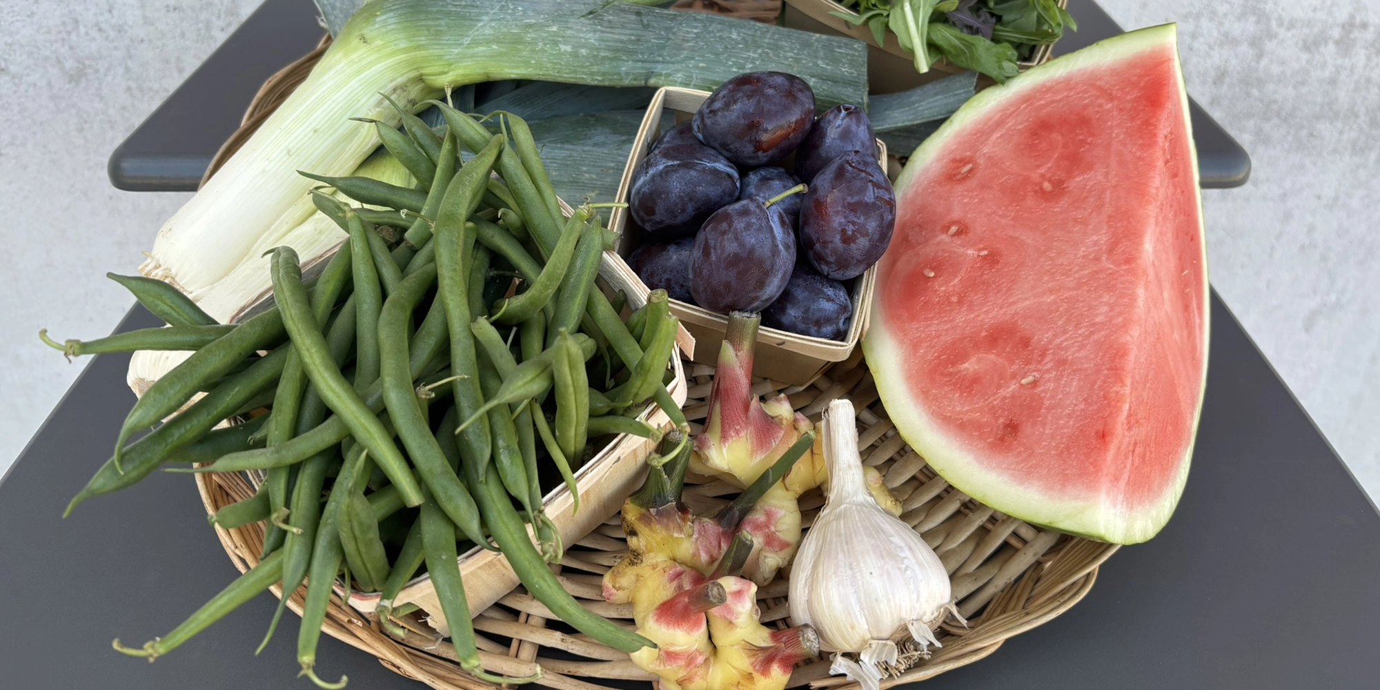 A variety of fruits and vegetables inside a wide wicker basket, including snap peas and watermelon