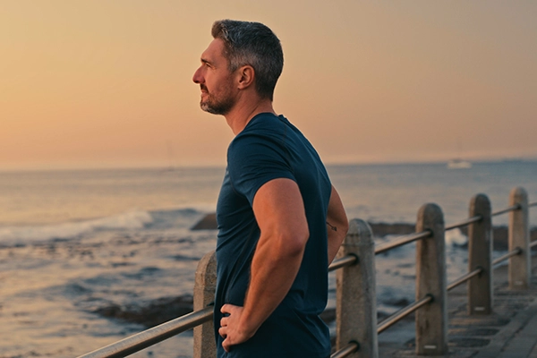 A middle-aged fit man stands on a pier with his hands on his hips, looking out at a large lake