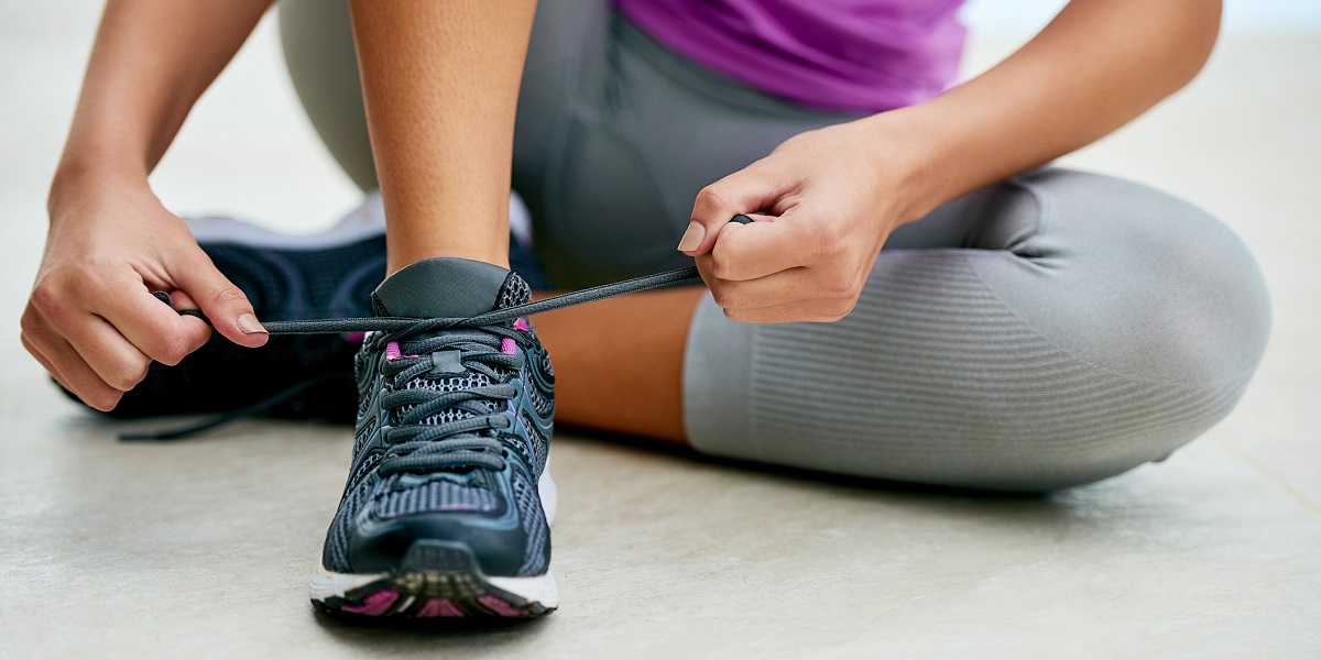 Woman wearing grey exercise pants and purple shirt is sitting on the floor, tying her right sneaker.