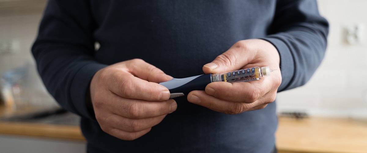 A man wearing a blue sweatshirt stands in his kitchen holding a syringe containing anti-obesity medication (AOM).