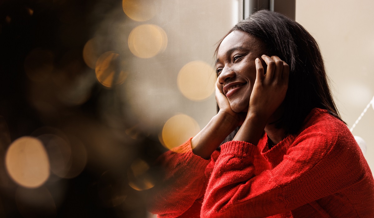 Young woman wearing red clothing, smiles while looking out the window with twinkling lights in the background.