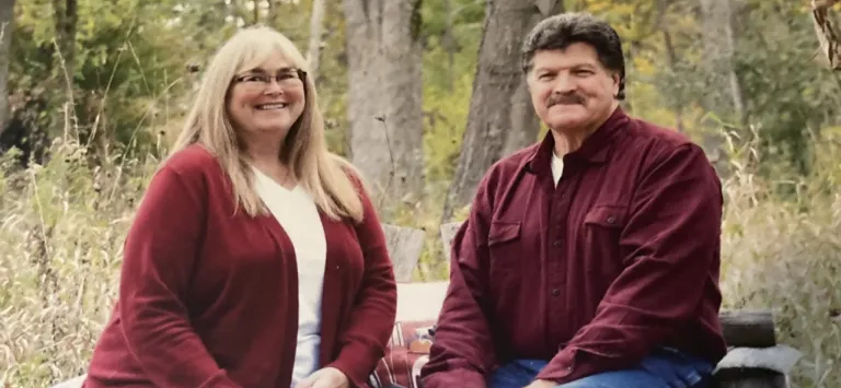 Caucasian woman and man smiling and sitting on a wood bench outside during fall in front of a group of trees.