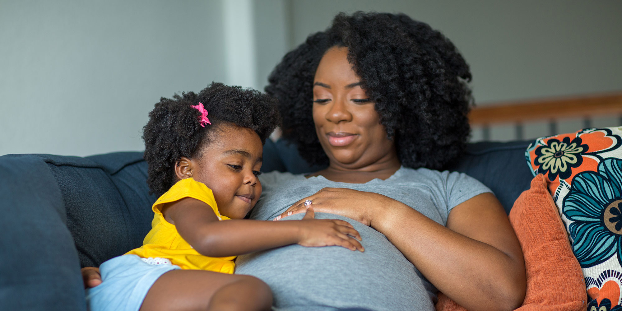 A pregnant woman shares a moment with her toddler daughter, who rests a hand on mom's pregnant belly